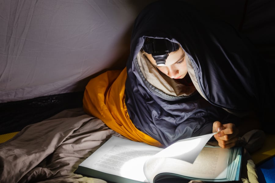 Child reading a book by headlamp in a tent.