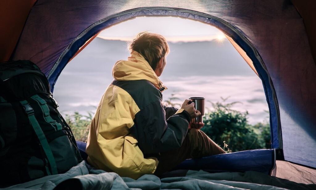 The Benefits of Solo Camping for Personal Growth 1 A young man sitting in the doorway of a tent with a stainless beverage mug, looking over a blurry landscape.