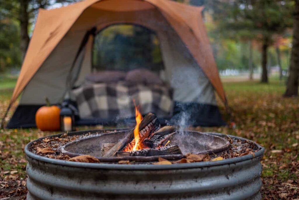 A cozy-looking campsite in the fall with an air matters bed inside a tent and a campfire near the tent.